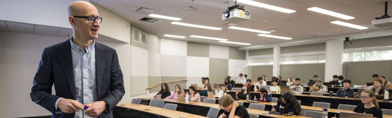 Economics classroom with professor at the front and students sitting at long desks listening and writing notes.