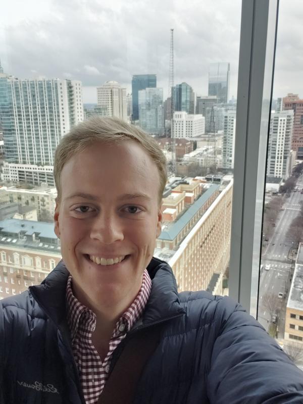 Selfie of Assistant Professor Dylan Brewer in front of a glass wall with a view of the Atlanta skyline