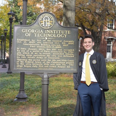 Image of Patrick in a graduation robe posing in front of the Georgia Tech sign